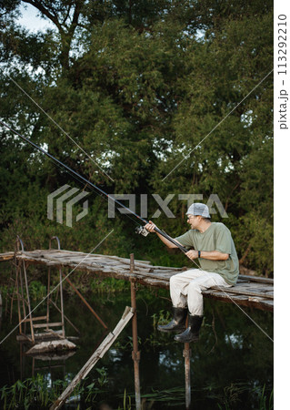 Man with fishing rod near the lake fishing at summer. Fisherman sitting on a footbridge. Man wearing khaki clothes and a cap. 113292210