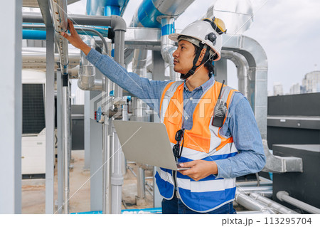 engineer man in a safety vest is standing around gas plump pipe, looking at a laptop. He is wearing a hard hat and a helmet engineer man in a safety vest is standing around gas plump pipe, looking at a laptop. He is wearing a hard hat and a helmet 113295704