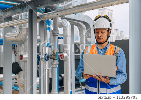 engineer man in a safety vest is standing around gas plump pipe, looking at a laptop. He is wearing a hard hat and a helmet engineer man in a safety vest is standing around gas plump pipe, looking at a laptop. He is wearing a hard hat and a helmet 113295705