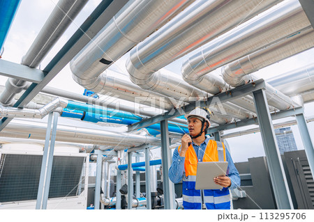 engineer man in a safety vest is standing around gas plump pipe, looking at a laptop. He is wearing a hard hat and a helmet engineer man in a safety vest is standing around gas plump pipe, looking at a laptop. He is wearing a hard hat and a helmet 113295706