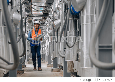 man in a safety vest is walking through a large industrial building with pipes and wires. He is holding a clipboard inspecting pipeline area. 113295707