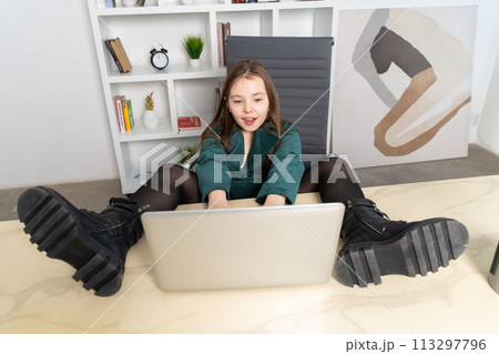 Little cheerful girl sitting with laptop with her feet on the table. High quality photo 113297796
