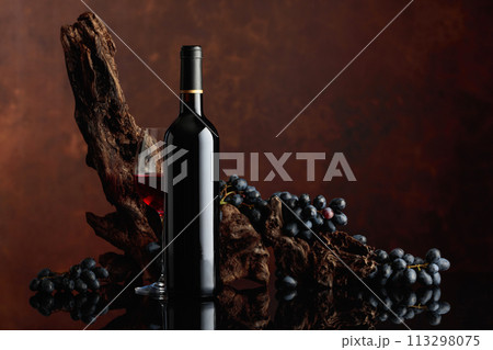 Bottle and glass of red wine with an old snag and blue grapes. 113298075