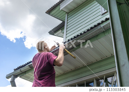Adult man, owner of dacha with nail and an axe replacing hammer examines roof for repair. Reparation with his own hands. Rear view 113303845