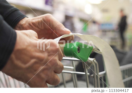 The hands of an unrecognizable older man hold a sales receipt next to a grocery cart in a supermarket. The hands of an unrecognizable older man hold a sales receipt next to a grocery cart in a supermarket. 113304417