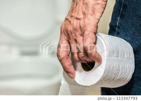 An older man's hand holding a roll of toilet paper in front of the toilet. An older man's hand holding a roll of toilet paper in front of the toilet. 113307793