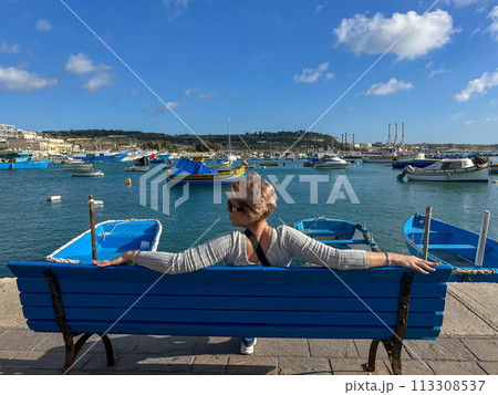 Woman sitting on bench and looking at boats in Marsaxlokk harbor, Malta 113308537