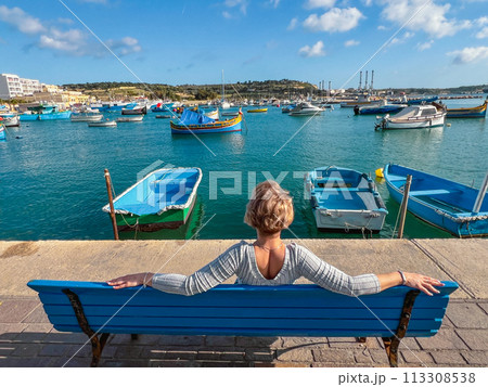 Woman sitting on bench and looking at boats in Marsaxlokk harbor, Malta 113308538