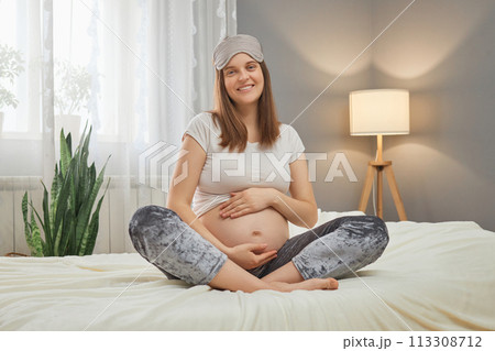 Attractive smiling cheerful Caucasian pregnant woman sitting in bed and holding her belly looking at camera with happy smile enjoying her pregnancy stroking tummy 113308712