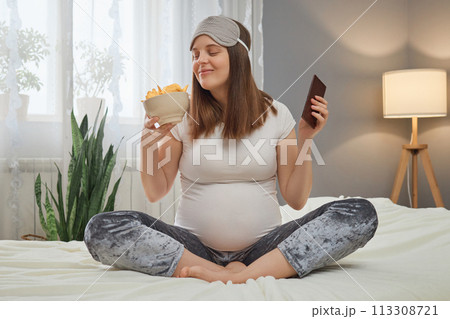 Funny smiling pregnant woman sitting in lotus pose holding bowl with potato chips and bar of chocolate smelling harmful fast food braking pregnant diet 113308721