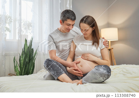 Beautiful dark haired pregnant woman and her handsome husband smiling while spending time together sitting in bed couple stroking belly Beautiful dark haired pregnant woman and her handsome husband smiling while spending time together sitting in bed couple stroking belly 113308740