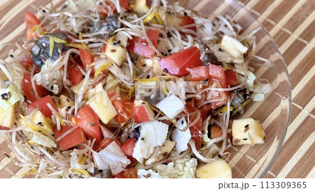 Salad of fresh vegetables and microgreens. Tomatoes, onions, apples, a mound of sprouted mung beans, sesame seeds, flax and cheese cubes on on a transparent plate on the table. Close-up 113309365