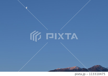 南アルプス鳳凰三山の山々が朝日に輝くとき空高く残る月齢22.2の月 113310475