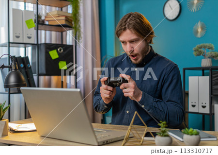 Happy freelancer business man playing video games on laptop holding controller at home office 113310717