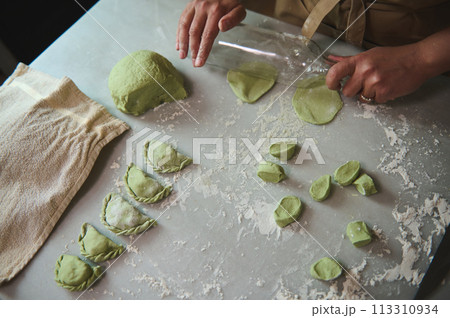 Top view woman's hands using a glass wine bottle for rolling out the dough with green spinach, cooking traditional Ukrainian Varennyky with mashed potatoes. Food. Traditions. Culture 113310934