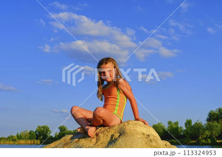 Adorable little girl sitting on a big stone in the lake on a hot summer day. Child dressed in swimsuit. Summer and happy childhood concept. Copy space in bright blue sky 113324953