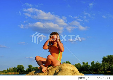 Beautiful girl sitting on a big rock. Little girl is wearing swimsuit. Girl is making heart shape gesture by her hands. Summer and happy childhood concept. Copy space in bright blue sky Beautiful girl sitting on a big rock. Little girl is wearing swimsuit. Girl is making heart shape gesture by her hands. Summer and happy childhood concept. Copy space in bright blue sky 113324954