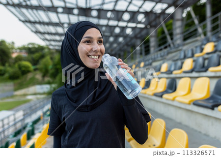 A Muslim woman in hijab holding a water bottle at a sports stadium, embodying health and fitness. 113326571
