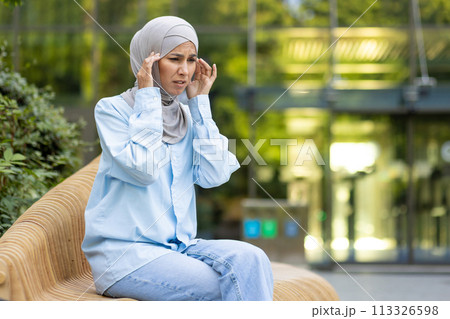 A woman wearing a hijab outdoors appears stressed and holds her head, showing signs of a headache or anxiety. 113326598