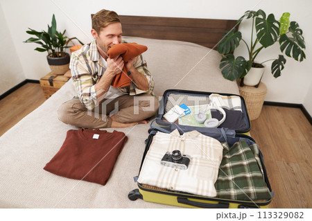 Portrait of happy young man, smells his clothes with pleased face, sits on bed with suitcase, goes on holiday, tries new detergent 113329082