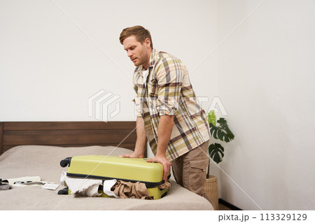 Portrait of young guy, traveller going on holiday, pressing his suitcase, trying to push clothes inside bag, closing his luggage, struggling to pack lots of things for vacation Portrait of young guy, traveller going on holiday, pressing his suitcase, trying to push clothes inside bag, closing his luggage, struggling to pack lots of things for vacation 113329129