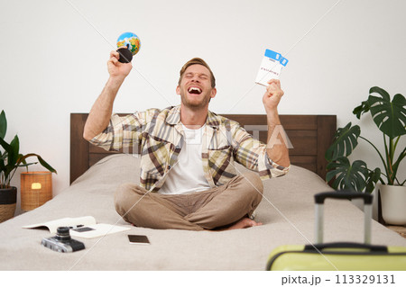 Portrait of happy guy, tourist goes on vacation abroad, holds globe and plane ticket, excited about his holiday, sits on bed with smiling, enthusiastic face expression Portrait of happy guy, tourist goes on vacation abroad, holds globe and plane ticket, excited about his holiday, sits on bed with smiling, enthusiastic face expression 113329131