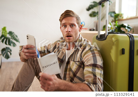 Man with scared face, sits on floor near suitcase, holds smartphone and passport Man with scared face, sits on floor near suitcase, holds smartphone and passport 113329141
