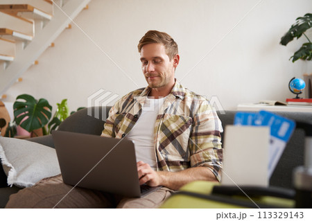 Portrait of handsome young man, has two tickets and passport, goes online on laptop, booking hotel and planning his holiday, sitting in living room Portrait of handsome young man, has two tickets and passport, goes online on laptop, booking hotel and planning his holiday, sitting in living room 113329143