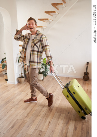 Full length photo of happy guy with suitcase, walking in apartment, shows phone call gesture 113329219