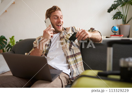 Image of guy calling someone, holding credit card and using laptop, has suitcase and camera packed for holiday Image of guy calling someone, holding credit card and using laptop, has suitcase and camera packed for holiday 113329229
