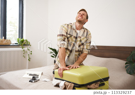 Portrait of young man sweating, shutting his suitcase, trying to close the luggage with lots of clothes, sitting on bed, packing things for summer vacation, going on holiday or business trip 113329237