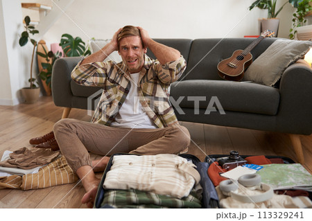 Portrait of worried man, tourist looking stressed and concerned, packing suitcase, holding hands on head, deciding on what to bring on holiday Portrait of worried man, tourist looking stressed and concerned, packing suitcase, holding hands on head, deciding on what to bring on holiday 113329241