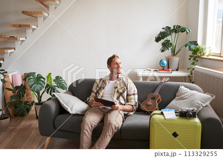 Portrait of handsome young man, tourist sitting with digital tablet near suitcase and two tickets, booking something online for his vacation, preparing for holiday 113329255