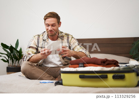 Portrait of handsome man, tourist, looking at his holiday tickets, booked vacation with travel agency, sitting on bed and packing clothes in suitcase 113329256