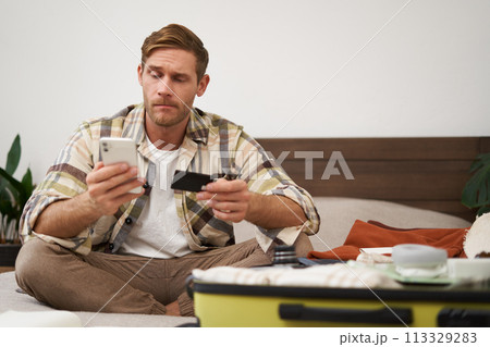 Angry man holds credit card and mobile phone, sits next to clothes in suitcase, has problems with paying online Angry man holds credit card and mobile phone, sits next to clothes in suitcase, has problems with paying online 113329283