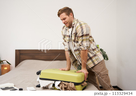 Portrait of young guy, traveller going on holiday, pressing his suitcase, trying to push clothes inside bag, closing his luggage, struggling to pack lots of things for vacation 113329434