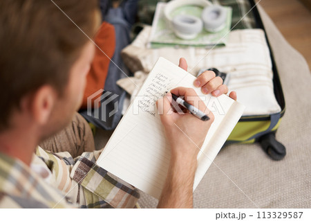 Close up portrait of man packing suitcase, making notes, check-list of items he wants to take on holiday, packing luggage 113329587