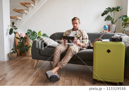 Image of young man with tablet, sitting in living room with suitcase, reading, studying the route, preparing for a vacation Image of young man with tablet, sitting in living room with suitcase, reading, studying the route, preparing for a vacation 113329588