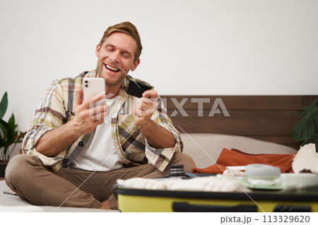 Cheerful tourist, guy with credit card and mobile phone, paying for holiday tickets, sitting on bed with suitcase, packing and going on holiday 113329620
