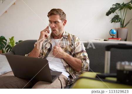 Portrait of angry man, looking frustrated, talking on mobile phone, sitting with laptop and suitcase 113329623