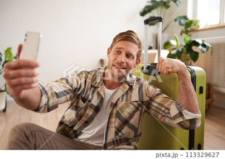 Image of handsome smiling young man going on holiday, posting online about vacation, taking selfie on smartphone with suitcase, shows muscles 113329627