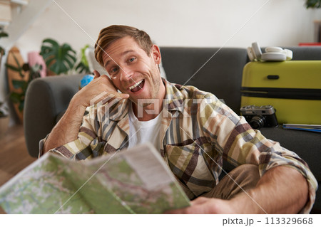 Portrait of handsome smiling young man, tourist shows call me gesture, phone hand, holding travel map, inviting to go on holiday, packed suitcase 113329668