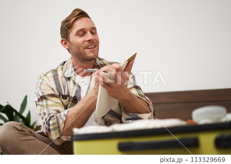 Portrait of thoughtful young man, tourist going on vacation, going through holiday check-list, writing down what he wants to take on travelling Portrait of thoughtful young man, tourist going on vacation, going through holiday check-list, writing down what he wants to take on travelling 113329669