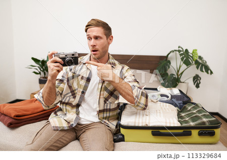 Portrait of handsome happy man, holding his analogue camera, sitting near opened suitcase, packing things for holiday, travelling abroad, getting ready for vacation Portrait of handsome happy man, holding his analogue camera, sitting near opened suitcase, packing things for holiday, travelling abroad, getting ready for vacation 113329684