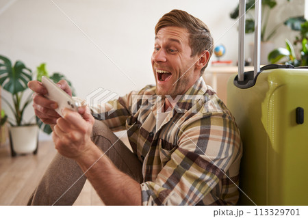 Happy man playing on mobile phone, laughing and smiling, sitting with suitcase, going on holiday, travelling Happy man playing on mobile phone, laughing and smiling, sitting with suitcase, going on holiday, travelling 113329701