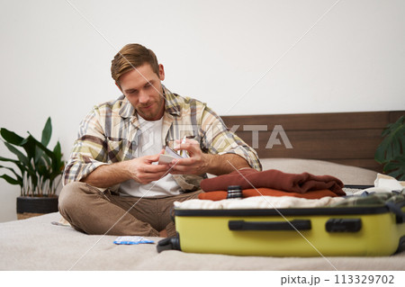 Portrait of young man with suitcase, holding his plane tickets, preparing to go on holiday, packing things for vacation, tourist puts clothes in bag Portrait of young man with suitcase, holding his plane tickets, preparing to go on holiday, packing things for vacation, tourist puts clothes in bag 113329702