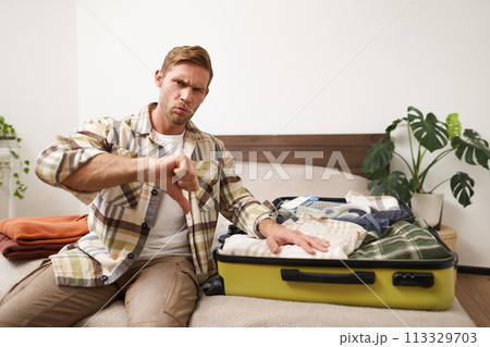 Image of young disappointed man, showing thumbs down, puts hand on his clothes inside luggage, dislike, disapprove something awful, upset with his holiday Image of young disappointed man, showing thumbs down, puts hand on his clothes inside luggage, dislike, disapprove something awful, upset with his holiday 113329703