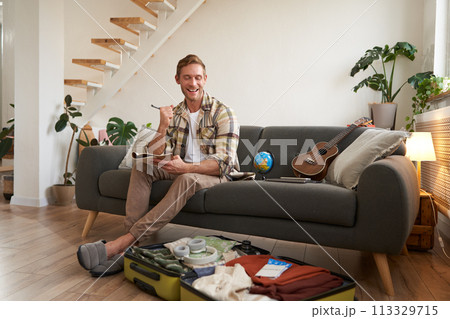 Happy man with pen and notebook, checking list of items and packing suitcase for journey, going on holiday, preparing for summer vacation, sitting in living room Happy man with pen and notebook, checking list of items and packing suitcase for journey, going on holiday, preparing for summer vacation, sitting in living room 113329715