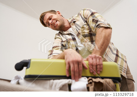 Side view of young man pushing on suitcase, trying to close it, has lots of clothes sticking out of luggage, going on long holiday, packing for vacation 113329718