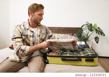 Tourism and travelling concept. Portrait of handsome young man going on holiday, packing his suitcase, checking luggage, sitting on bed and looking at packed things Tourism and travelling concept. Portrait of handsome young man going on holiday, packing his suitcase, checking luggage, sitting on bed and looking at packed things 113329721